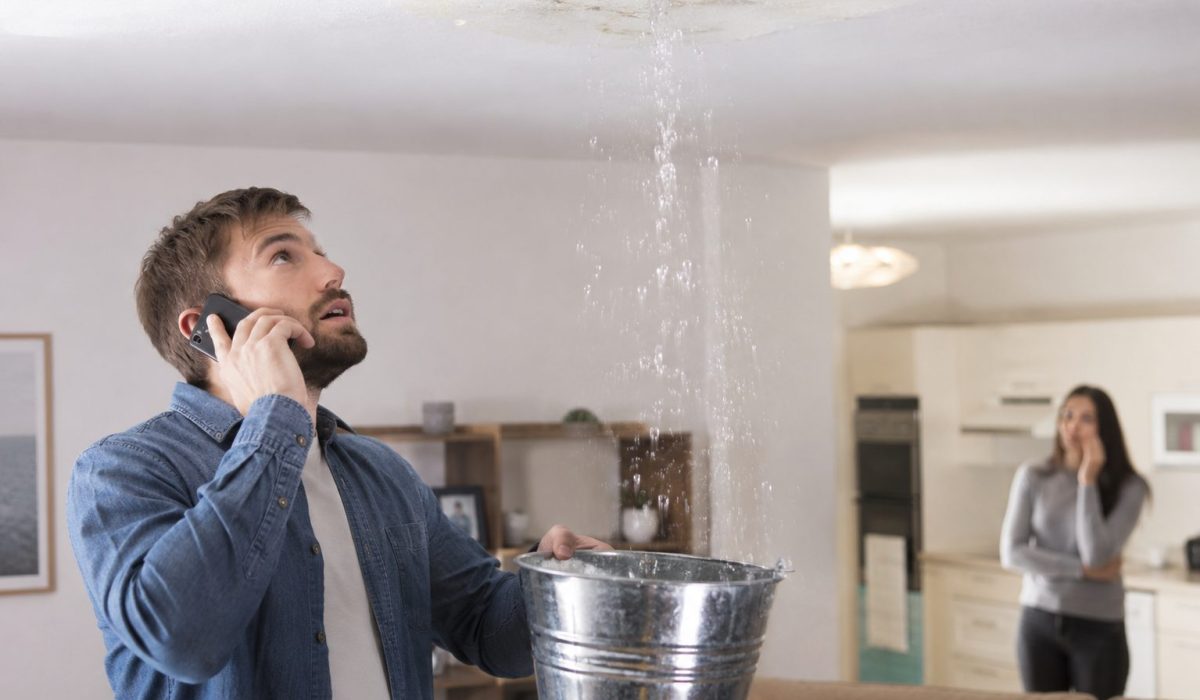 Homme appelant son assurance pendant qu’une fuite d’eau au plafond est recueillie dans un seau après un dégât des eaux