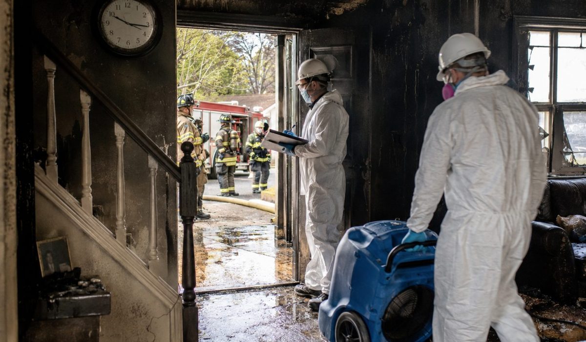 Professionnels en combinaison de protection intervenant dans une maison endommagée par un incendie avec pompiers, suie, eau d’extinction et matériel de séchage
